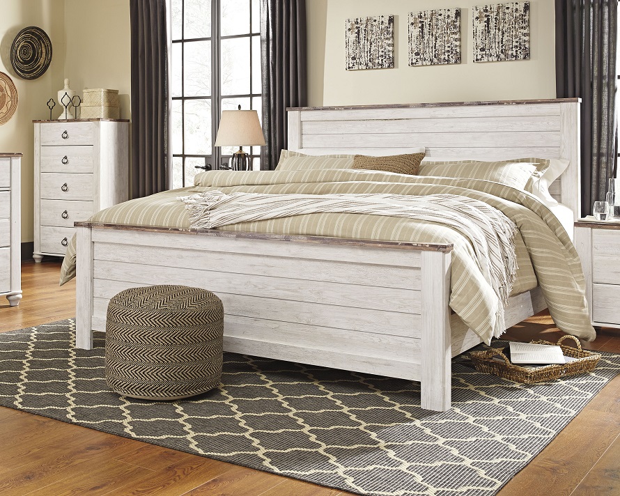 White coastal bed with distressed wood with tan bedding a geometric rug and a pouf on the rug.